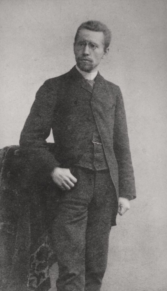 Die historische schwarz-weiß-Aufnahme zeigt Karl Kollwitz um 1885. Er ist in stehender Position. Er trägt eine Brille und einen Ziegenbart. Er trägt ein dreiteiligen dunklen Anzug. Sein rechter Daumen ist in seine rechte Hosentasche eingehakt. Seine linke Hand hat er zu einer leichten Faust geballt. Sein Blick ist auf einen Punkt außerhalb der Kamera gerichtet.