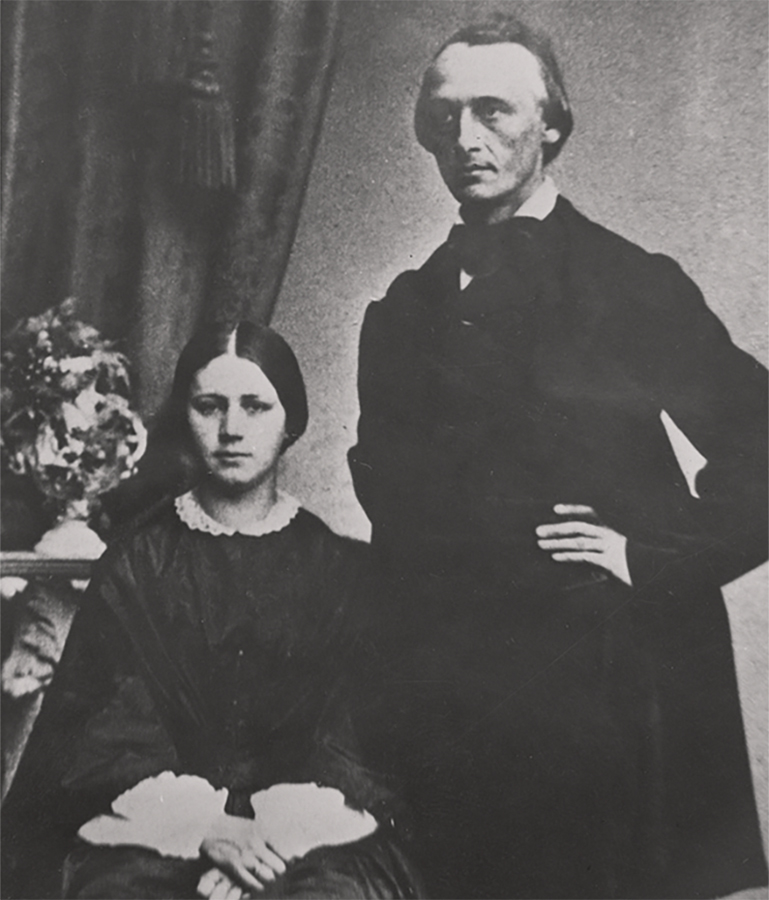 Die historische schwarz-weiß-Aufnahme zeigt das Ehepaar Schmidt. Auf dem Doppelporträt tragen sie dunkle Kleidung. Katharina sitzt und blickt direkt in die Kamera; Carl steht, mit den Händen in den Hüften, und blickt auf einen Punkt außerhalb der Kamera.
