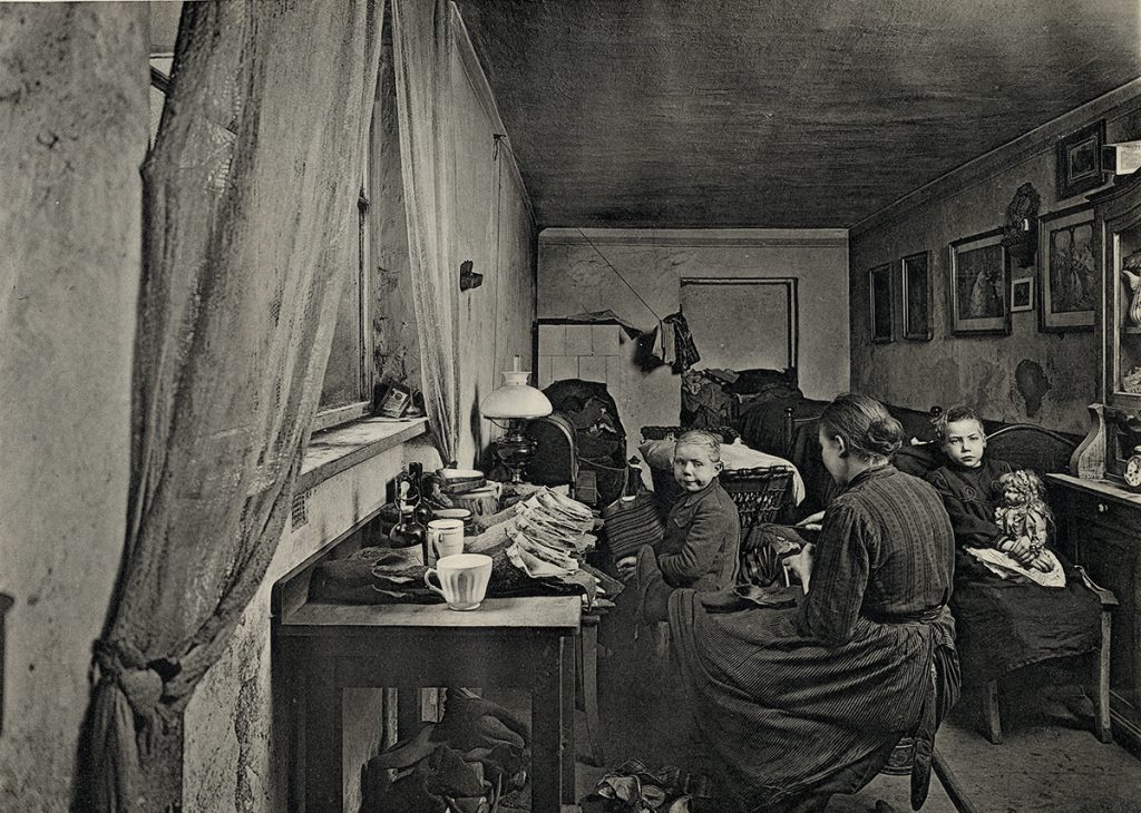 Eine historische Aufnahme zeigt eine Heimarbeiterin mit ihren Kindern in einer Kellerwohnung in der Rheinsbergerstraße 62 im Jahre 1906.