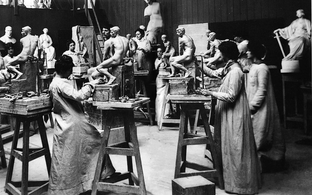 Historische Fotografie von Schülerinnen einer Bildhauerklasse in Arbeitskleidung. Sie stehen in einem Atelierraum voller Skulpturen. Die Frauen sind konzentriert am Arbeiten. Als Vorbild dient ihnen ein männliches Aktmodell.