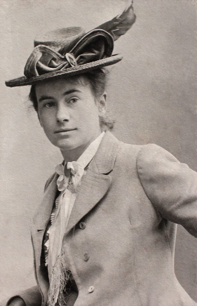 Historische Porträtaufnahme der jungen Marianne Fiedler um 1890. Man sieht sie als Halbﬁgur, die den Betrachter anblickt. Sie trägt zeittypische Kleidung: Ein Jackett, ein Halstuch mit einer Brosche und einen auﬀälligen Hut mit Schleife und Feder verziert.