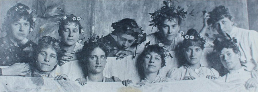 Historische Fotografie der Münchner Damenklasse. Zehn jungen Frauen, die Blumenkränze auf ihren Köpfen tragen. Sie posieren gemeinsam für ein Gruppenfoto. Sie spannen ein weißes Tuch. Man sieht größtenteils nur ihre Köpfe. Käthe Schmidt befindet sich in der Bildmitte, in der hinteren Reihe. Sie hat ihren Blick von der Kamera abgewandt. Ihre Hände sind ineinander gefaltet.