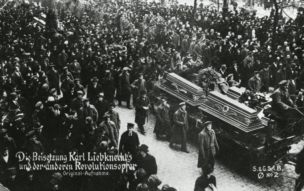 Eine historische Fotografie zeigt den Trauerzug für Karl Liebknecht und die anderen Revolutionsopfer. Die Särge werden auf Kutschen transportiert, umringt von Meschen, die dem Trauerzug folgen.