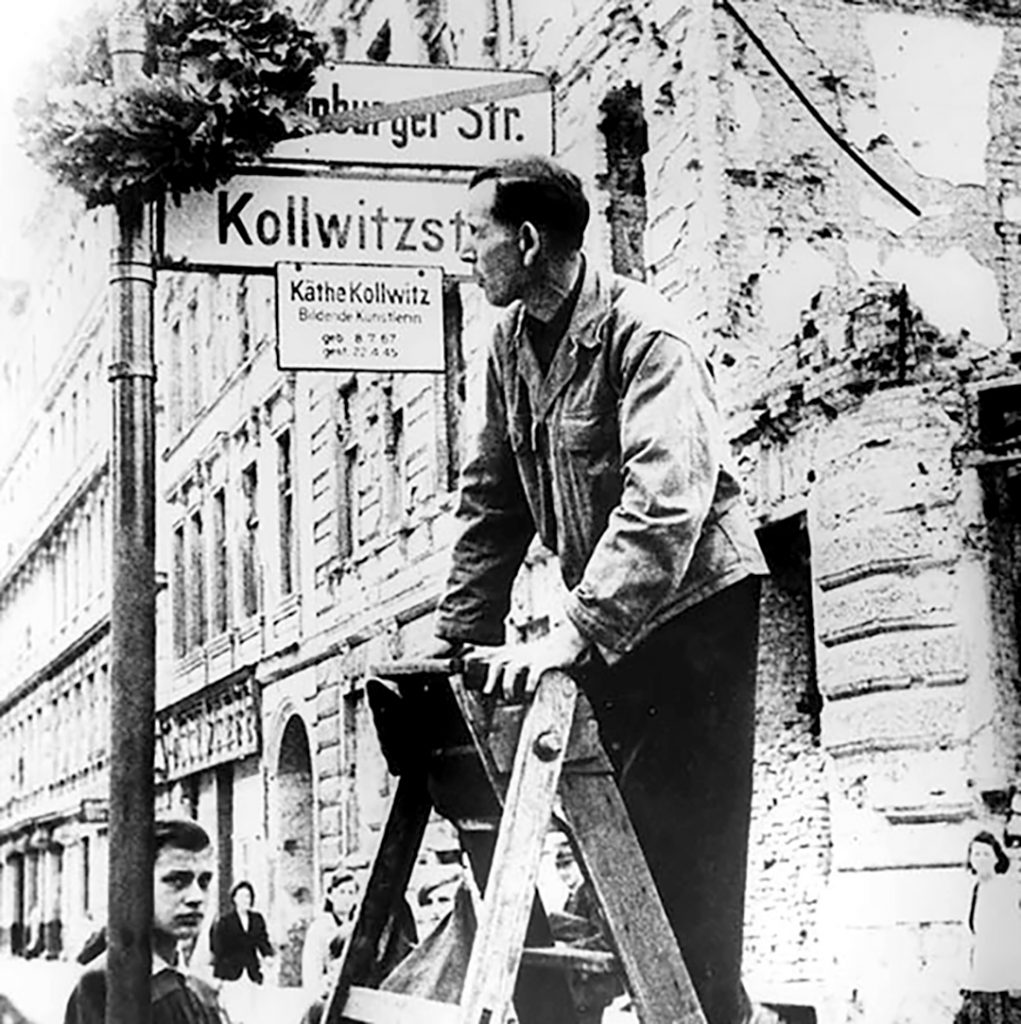 Historische Fotografie, die eine Straßenumbenennung dokumentiert. Zwei Männer stehen im Vordergrund. Einer der beiden Männer steht auf einer Leiter und hat das neue Straßenschild angebracht. Er scheint noch einen prüfenden Blick darauf zu werfen. Der andere Mann steht neben der Leiter und schaut direkt in die Kamera. Ein paar Passanten laufen an ihnen vorbei.