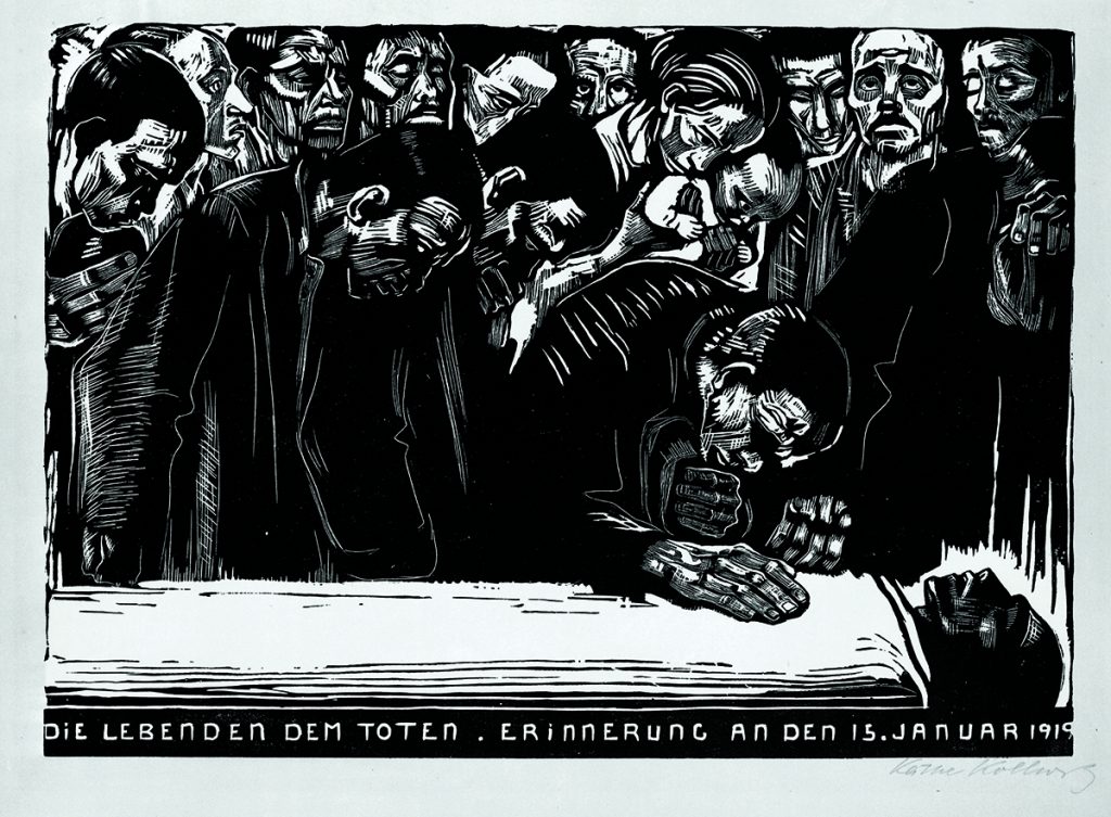 Ein aufgebahrter Leichnam. Um ihn herum steht eine Gruppe von Menschen, die seinen Tod betrauern. Ein Mann steht unmittelbar neben ihm. Er ist zu ihm heruntergebeugt und hat ihm seine rechte Hand auf die Brust gelegt. Unter den größtenteils männlichen Trauernden befindet sich auch eine Frau mit Säugling auf dem Arm in der Bildmitte. Am unteren Bildrand steht in großen Lettern: Die Lebenden dem Toten. Erinnerung an den 15. Januar 1919. Rechts unten signiert: Käthe Kollwitz
