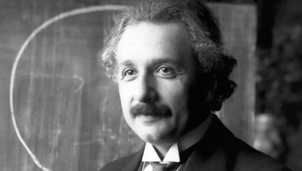 Die historische Aufnahme zeigt Albert Einstein während eines Vortrags in Wien im Jahr 1921. Er trägt einen Anzug und steht lächelnd vor einer Tafel.