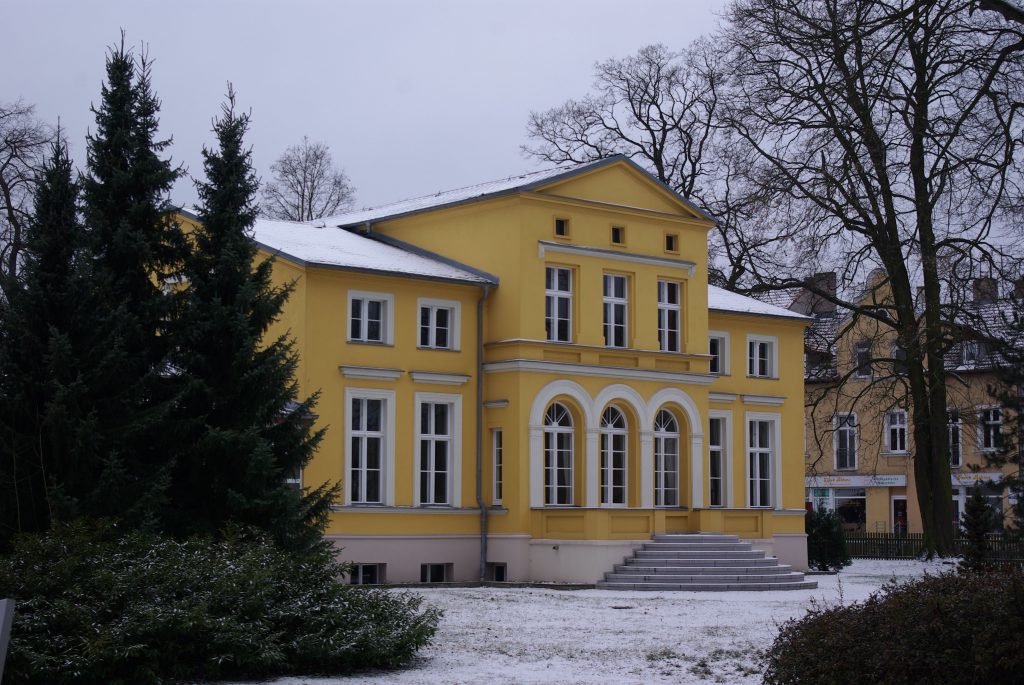 Fotograﬁe vom Gerhart-Hauptmann-Museum in Erkner in der Villa Lassen. Die Hausfassade ist in einem gelben Ton gehalten. Die Fotograﬁe entstand an einem Wintertag. Die Dächer und der Boden sind schneebedeckt. Zum Haus führt eine halbrunde Treppenanlage, die mittig vor der Fassade angelegt ist.