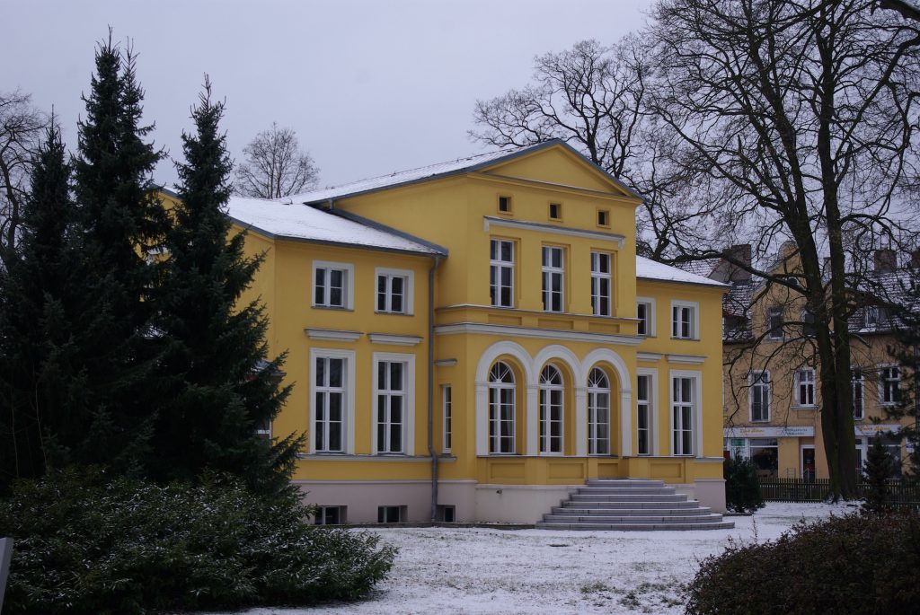 Fotografie vom Gerhart-Hauptmann-Museum in Erkner in der Villa Lassen. Die Hausfassade ist in einem gelben Ton gehalten. Die Fotografie entstand an einem Wintertag. Die Dächer und der Boden sind schneebedeckt. Zum Haus führt eine halbrunde Treppenanlage, die mittig vor der Fassade angelegt ist.