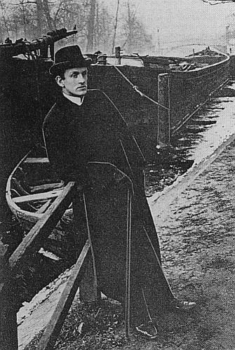 Historische Fotografie von Gerhart Hauptmann aus dem Jahr 1892. Ganzfigürlich dargestellt, an einem Schiffs-Kai stehend, dahinter Kähne. Er ist dunkel gekleidet mit Hut und Spazierstock ausgerüstet.