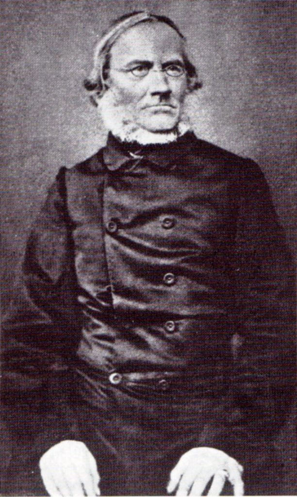 Die historische schwarz-weiß-Aufnahme zeigt Julius Rupp (1809-1884) als Halbfigur. Auf dem Porträt trägt er einen dunklen Anzug, einen Zweireiher, eine Brille und einen weißen Bart. Seine Arme sind angewinkelt; seine Hände ruhen auf den Oberschenkeln. 