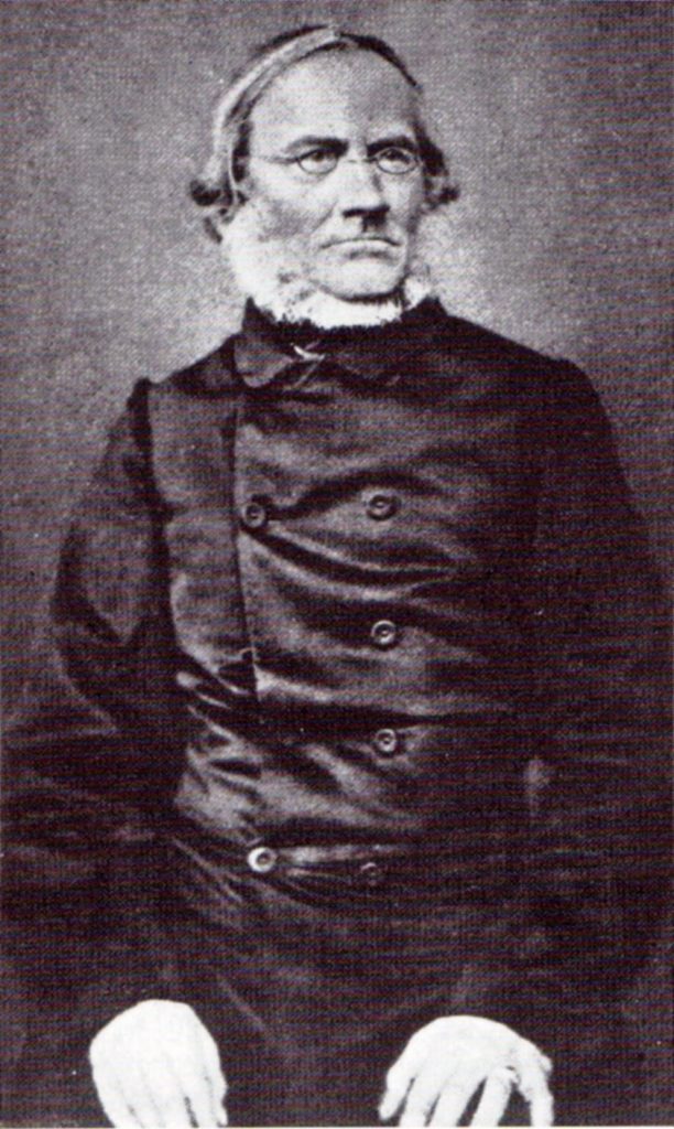 Die historische schwarz-weiß-Aufnahme zeigt Julius Rupp (1809-1884) als Halbfigur. Auf dem Porträt trägt er einen dunklen Anzug, einen Zweireiher, eine Brille und einen weißen Bart. Seine Arme sind angewinkelt; seine Hände ruhen auf den Oberschenkeln.