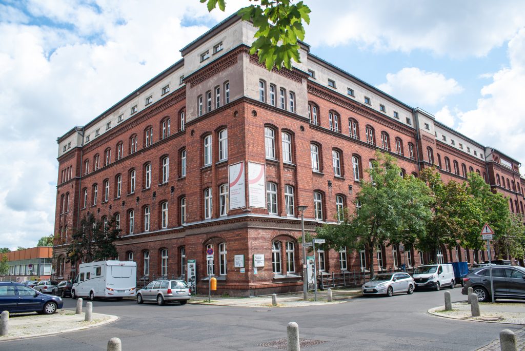 Auf dem Farbfoto ist die Fassade vor einem roter Backsteingebäude aus dem gegebüberliegenden Straßenkreuzung fotografiert zu sehen. Einige Autos parken davor.