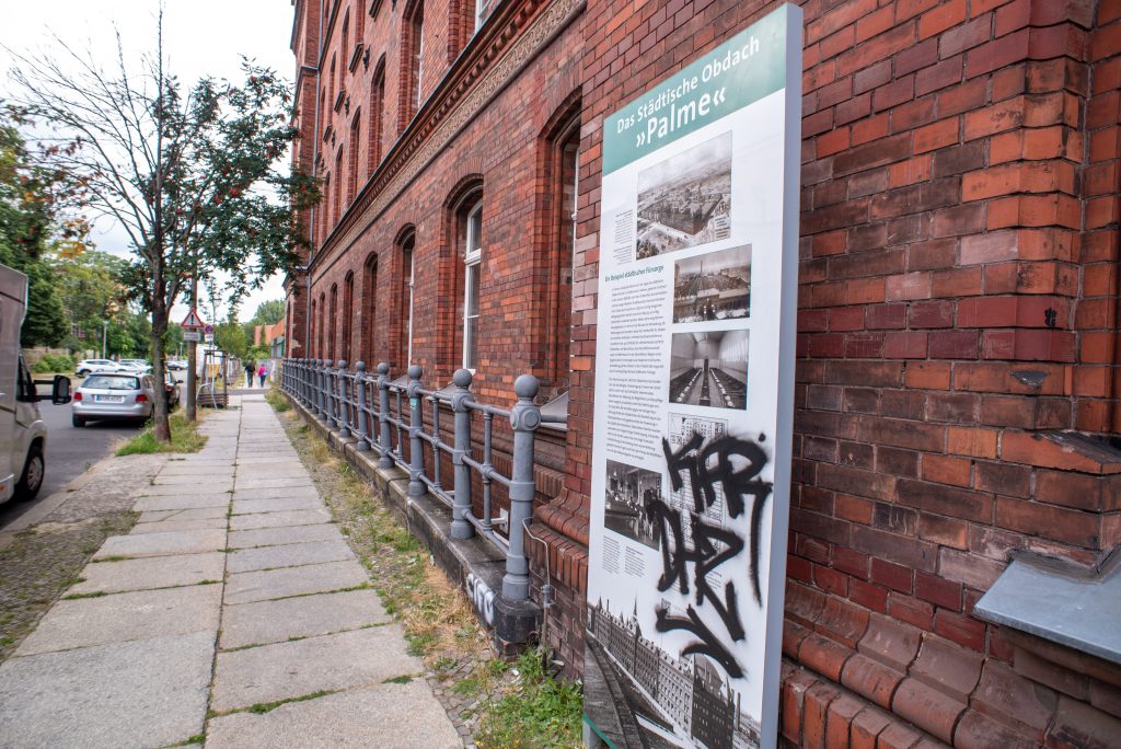 Auf dem Farbfoto ist seitlich die Fassade vor einem roter Backsteingebäude und einen Straßenausschnitt zu sehen. Vor dem Gebeude steht die Gedenktafel zum Obdachlosenasyl "Palme".
