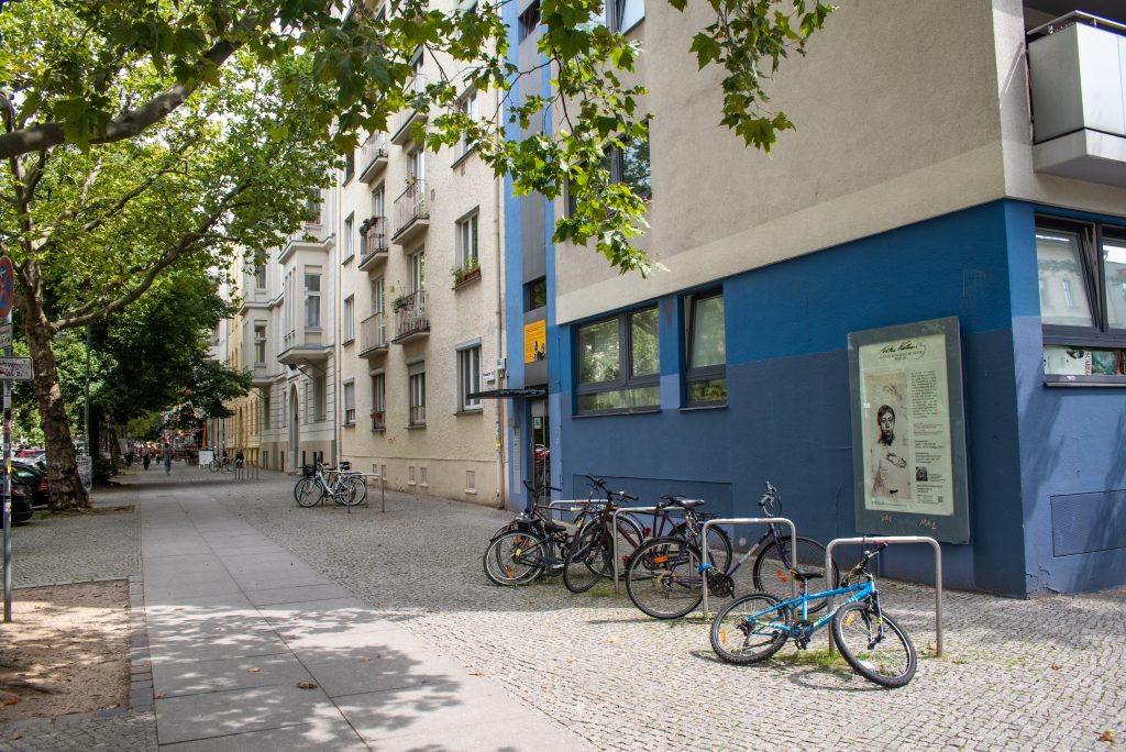 Eine Fotografie aus dem Jahr 2021 vom Kollwitzplatz. Einen Straßenausschnitt, die Fassade des Wohnhauses in der Kollwitzstr. 58 und die Gedenktafel zu Käthe Kollwitz ist auf dem Bild zu sehen. Einige Fahhreder parken vor dem Haus.