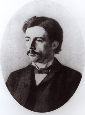 Die historische schwarz-weiß-Aufnahme zeigt Konrad Schmidt um 1885. Er trägt einen Schnurrbart, ein weißes Hemd mit Fliege, eine dunkle Weste und einen dunklen Mantel. Er schaut zur Seite, sein Blick ist auf einen Punkt außerhalb der Kamera gerichtet.